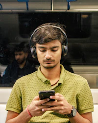 A young man wearing over-ear headphones looking at his phone while seated on a metro train
