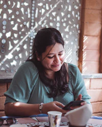 A smiling woman at a café table looking at her phone, with a paper cup in front of her