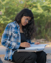 A young woman in glasses sitting outdoors with an open notebook on her lap, studying