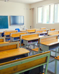An empty classroom with wooden desks, a green chalkboard and a wall-mounted screen